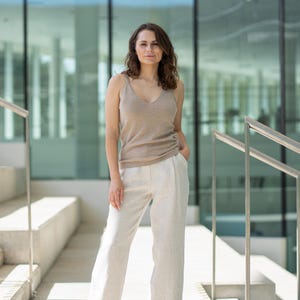 A woman with shoulder-length wavy brown hair stands looking directly at the camera, with one hand in her pocket and the other relaxed at her side. She is wearing a merino wool and cashmere tank top and linen menique wide-legged trousers, and is positioned on light-colored stairs with silver handrails and a modern building with large windows in the background.