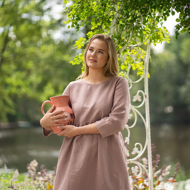 May include: A dusty rose-colored dress with three-quarter length sleeves and a round neckline, featuring a gathered waistline. The dress is held alongside a peach-colored ceramic pitcher decorated with a white floral pattern on its side.