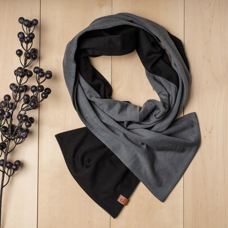 May include: A gray and black infinity scarf made of soft fabric. The scarf is folded in half and has a small brown leather tag with a letter "C" on it.