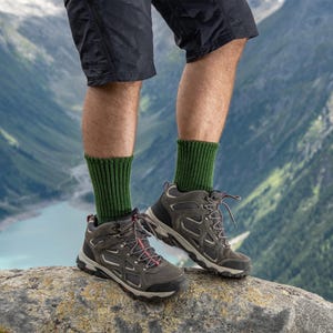 May include: A person wearing dark green hiking socks and gray hiking boots with red laces. The boots are on a rock with a mountain and lake background. The person is wearing black shorts.