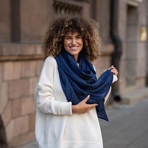 May include: A woman with curly brown hair is wearing a white sweater and a navy blue scarf. She is smiling at the camera.