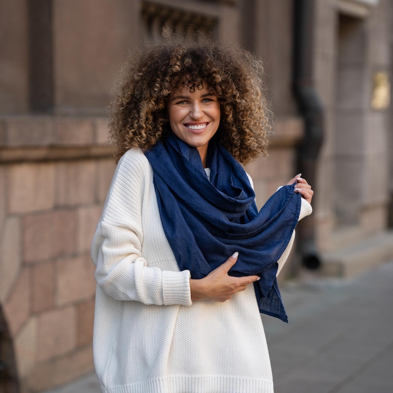 May include: A woman with curly brown hair is wearing a white sweater and a navy blue scarf. She is smiling at the camera.