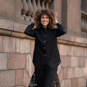May include: A woman with curly hair is wearing a black long-sleeved shirt with a collared neckline and button closure. She is standing in front of a stone wall.