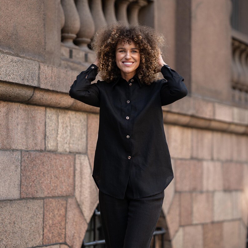 May include: A woman with curly hair is wearing a black long-sleeved shirt with a collared neckline and button closure. She is standing in front of a stone wall.