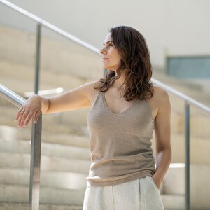 A woman with shoulder-length wavy brown hair stands in profile, looking to her left with one arm resting on a silver handrail and the other hand in her pocket. She is wearing a menique tank top and linen pants, and is positioned on light-colored stairs with a glass or clear barrier visible in the background.
