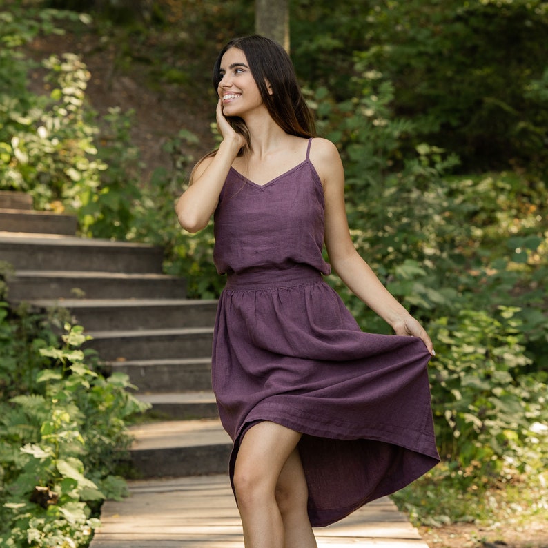 The model is wearing the SOPHIA skirt in shadow purple. She is looking sideways, with her hand on her chin, the other holding a side of her skirt. The skirt has a wide waist, and is matched with a same color slip top, tucked inside the skirt.