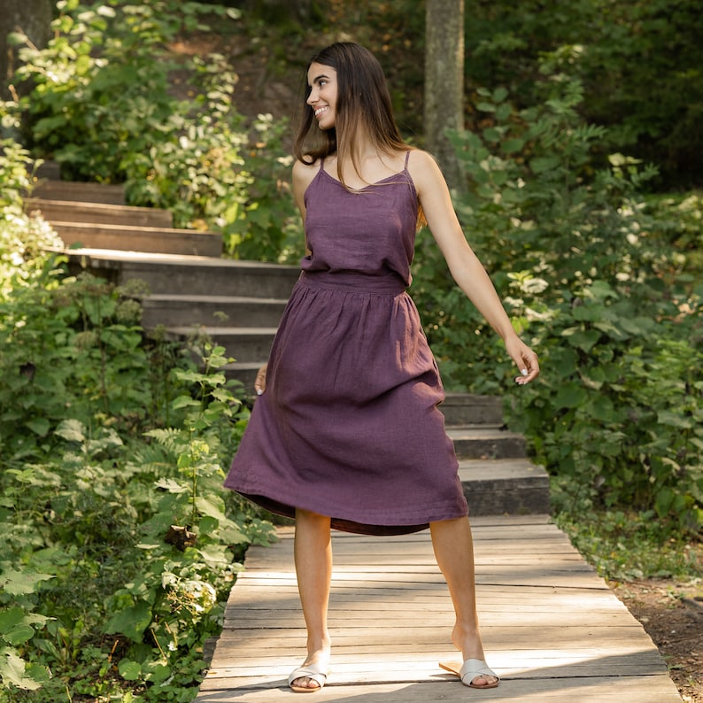 The model is wearing the SOPHIA skirt in shadow purple. She is outside, in a forest, on a wooden pathway. She is holding one side of the skirt, looking sideways, smiling. The skirt has a delicate A-line shape and a wide waist.
