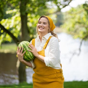 A cheerful woman stands near a tranquil lake, cradling a whole watermelon in her arms and smiling thoughtfully into the distance. She is styled in a pure white linen shirt layered under a spicy yellow jumpsuit with a coordinating bandana.