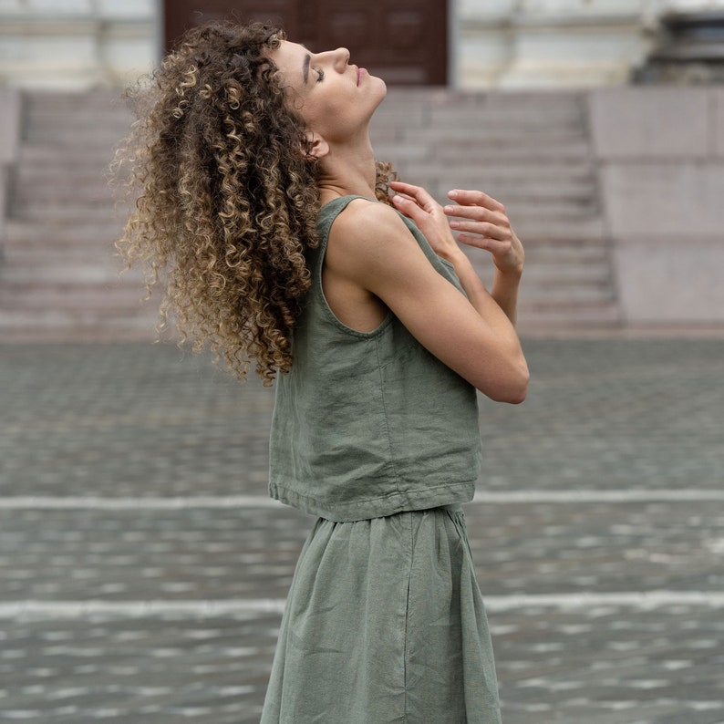 May include: A woman wearing a sleeveless linen top and skirt set in a muted green color. The top has a cropped, boxy fit and the skirt is a-line. The woman is standing with her head tilted back and her eyes closed.