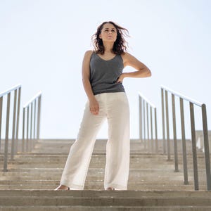 A woman with wavy brown hair stands confidently on outdoor concrete steps, looking slightly upwards with one hand on her hip. She is wearing a dark gray tank top and menique linen white trousers, with metal railings on either side of the stairs leading up to a bright sky.