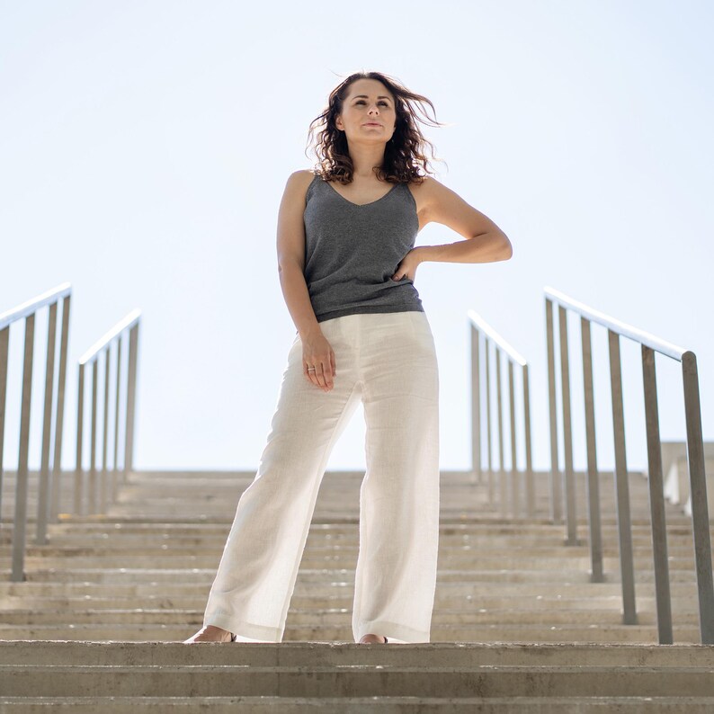 A woman with wavy brown hair stands confidently on outdoor concrete steps, looking slightly upwards with one hand on her hip. She is wearing a dark gray tank top and menique linen white trousers, with metal railings on either side of the stairs leading up to a bright sky.