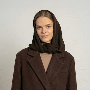 Puede incluir: Una mujer con una capucha de punto marrón oscuro y una blazer marrón a juego. La capucha está envuelta alrededor del cuello y la cabeza, creando un look cálido y elegante. La blazer tiene un diseño clásico con solapa de muesca.