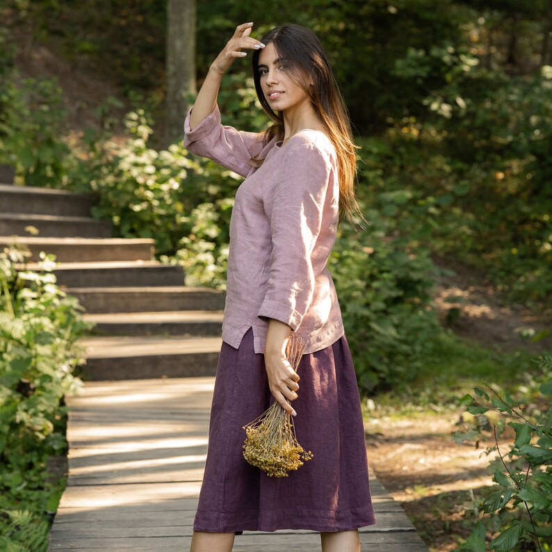 The model is wearing the SOPHIA skirt in shadow purple. She has her back facing the camera, holding a bouquet of wildflowers in her hands by her side. She is facing the camera, wearing a faded rose color long sleeve blouse, covering the waistline.