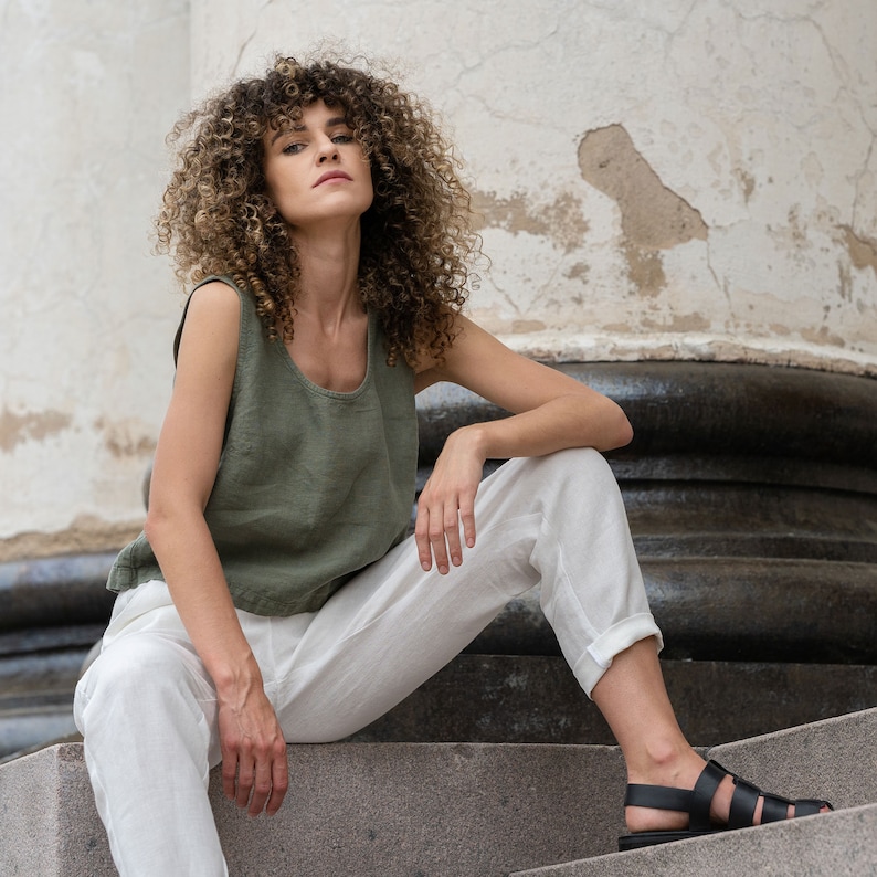 The model is wearing a linen clothing set, which includes a sleeveless crop top in stone green color and white high-waisted pants with pockets. She is sitting on stairs, with one leg higher than the other, hand resting on it.
