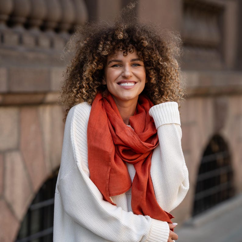 May include: A woman with curly brown hair is wearing a white sweater and a long, rust-colored scarf. She is smiling at the camera.