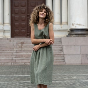 May include: A woman with curly brown hair is wearing a sleeveless, sage green linen dress. She is standing in front of a building with a brown door and white columns.
