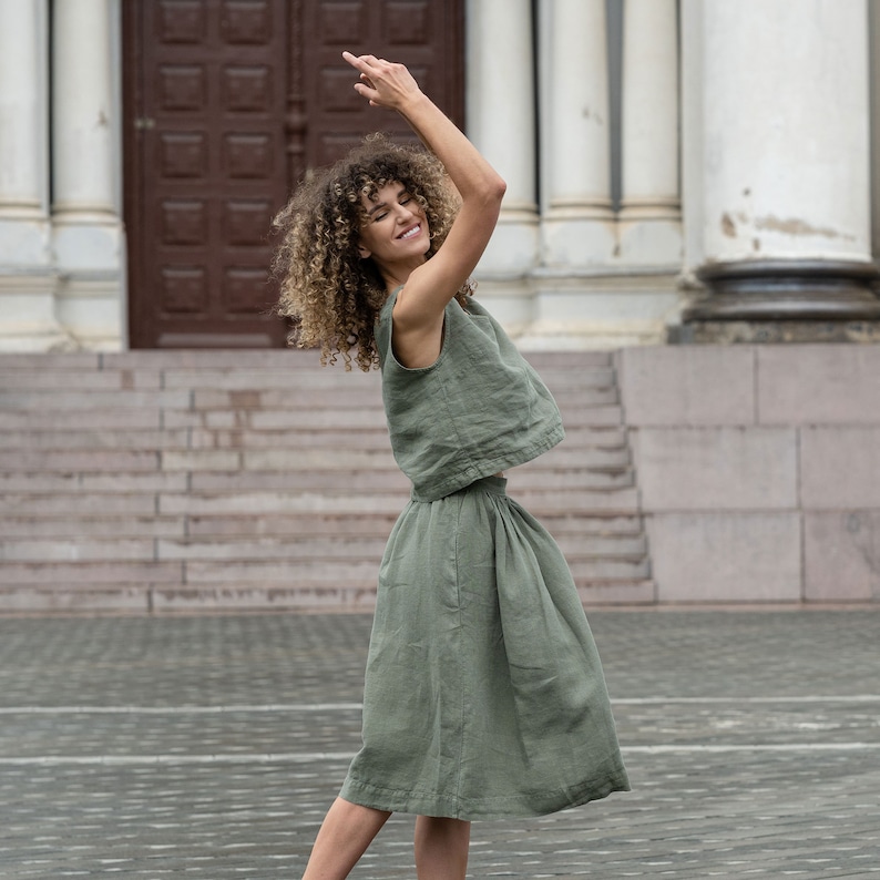 The model is wearing the SOPHIA skirt in stone green. She stands sideways, with both hands in the air above her head, smiling. The raised blouse reveals the fron of the waistline, which is wide. The skirt has a midi length.
