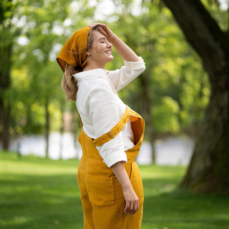 A smiling woman stands in a lush green park, turning her face toward the sun with her eyes closed while gently touching her head. She is wearing a pure white linen shirt layered under a vibrant, spicy yellow linen jumpsuit, accessorized with a matching yellow bandana.