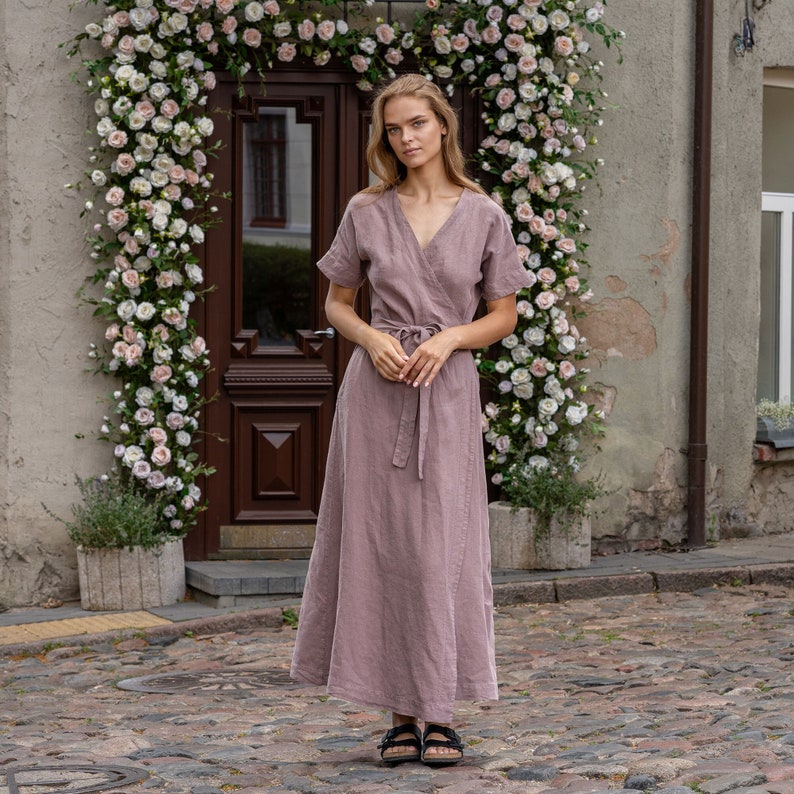 The model wears the ELEANOR maxi wrap dress in faded rose. She is standing full height, with her hands together, accentuating the waistline. The wrap design features a v-neck and a belt around the waist.