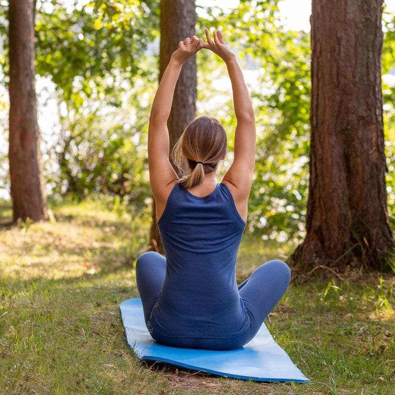 K&ouml;nnte beinhalten: Eine Frau in einem blauen Tanktop und einer blauen Hose sitzt auf einer blauen Yogamatte mit erhobenen Armen &uuml;ber dem Kopf. Sie praktiziert Yoga in einer Waldumgebung.