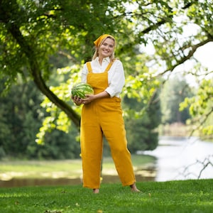 A smiling, barefoot woman stands on lush green grass, holding a whole watermelon at her waist. She is dressed in a vibrant spicy yellow linen jumpsuit over a pure white shirt, accessorized with a matching yellow bandana.