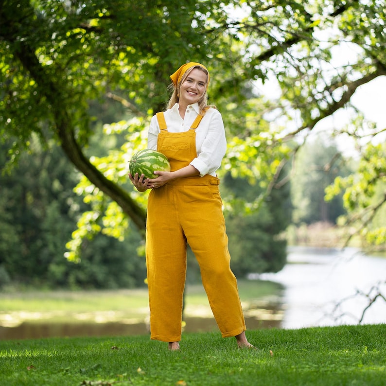 A smiling, barefoot woman stands on lush green grass, holding a whole watermelon at her waist. She is dressed in a vibrant spicy yellow linen jumpsuit over a pure white shirt, accessorized with a matching yellow bandana.