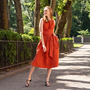 May include: A rust-colored linen dress with a self-tie sash and a sleeveless design. The dress is knee-length and has a relaxed fit.