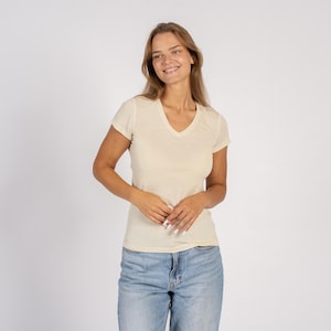 May include: A cream-coloured, short-sleeved V-neck t-shirt. The shirt is made of a soft material and is worn with blue jeans. The model is smiling and looking to the side.