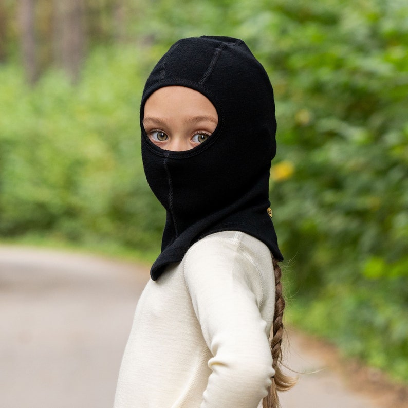 May include: A black balaclava with eye holes covers the head of a person wearing a white sweater. The person has long brown hair.