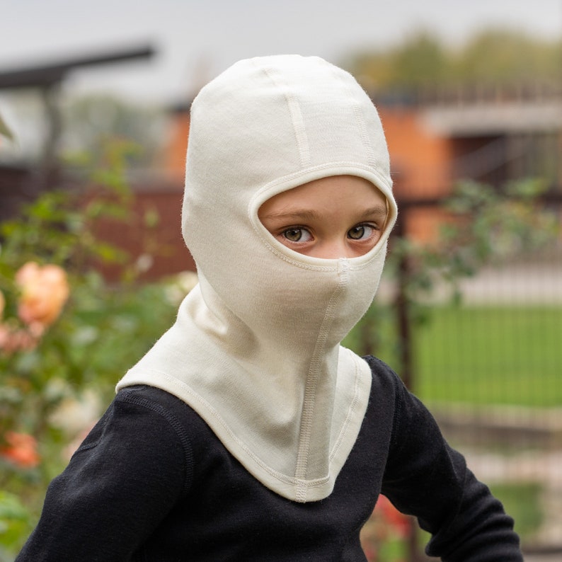May include: A white balaclava with a fold-over design covers the head and face of a child. The balaclava is made of a soft, knitted fabric. The child is wearing a black shirt.