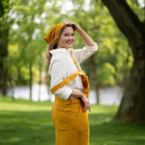 A woman stands in a sunlit green park, glancing happily over her shoulder directly at the camera with one hand resting gently on her head. She is dressed comfortably in a pure white linen shirt paired with a spicy yellow linen jumpsuit and a matching yellow bandana.