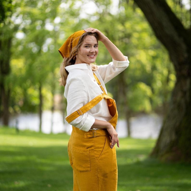 A woman stands in a sunlit green park, glancing happily over her shoulder directly at the camera with one hand resting gently on her head. She is dressed comfortably in a pure white linen shirt paired with a spicy yellow linen jumpsuit and a matching yellow bandana.
