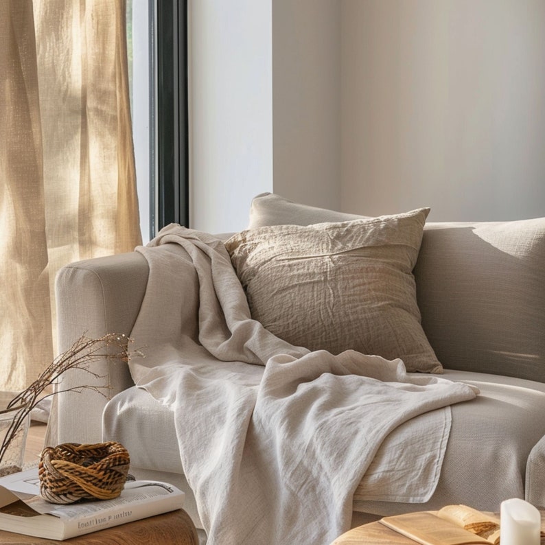 Pu&ograve; includere: Una coperta in lino beige chiaro drappeggiata su un divano con un cuscino in lino abbinato. Il divano si trova in un soggiorno moderno con una grande finestra e luce naturale.