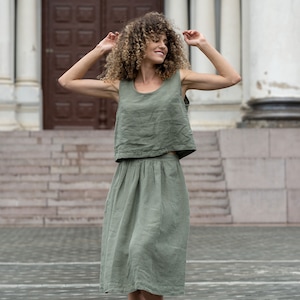The model is wearing the SOPHIA skirt in stone green. She has her hands bent at the elbow, raised to her shoulders, with her head turned to the side. She is smiling. The skirt is matched with a sleeveless blouse in the same color.