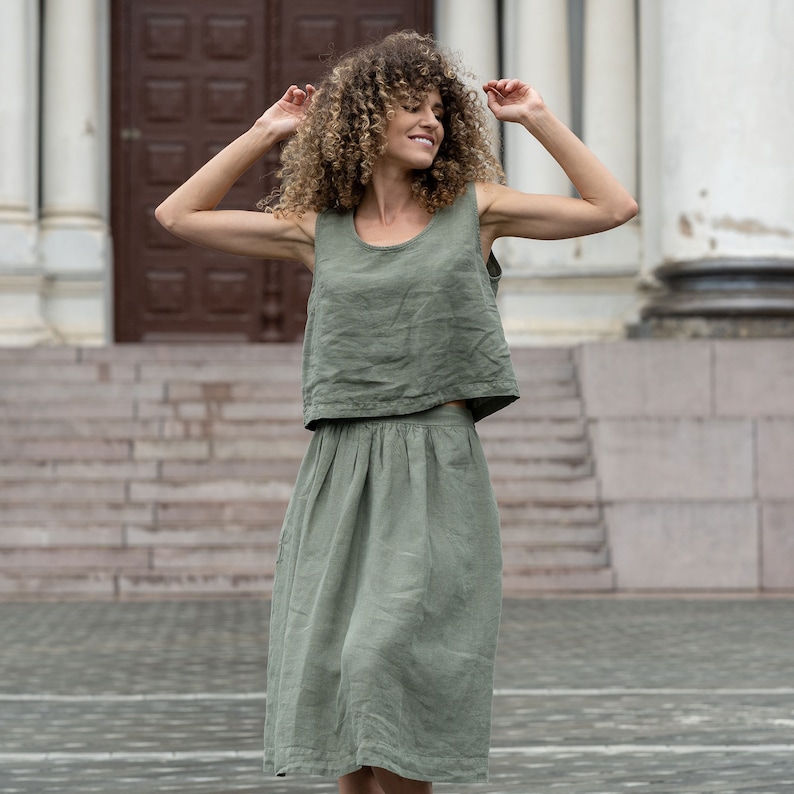 The model is wearing the SOPHIA skirt in stone green. She has her hands bent at the elbow, raised to her shoulders, with her head turned to the side. She is smiling. The skirt is matched with a sleeveless blouse in the same color.