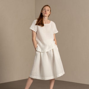 The model is wearing the SOPHIA skirt in pure white. She has her hands inside both concealed pockets. The skirt is midi-length, has an A-line shape.