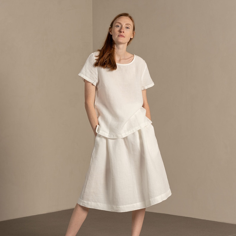The model is wearing the SOPHIA skirt in pure white. She has her hands inside both concealed pockets. The skirt is midi-length, has an A-line shape.
