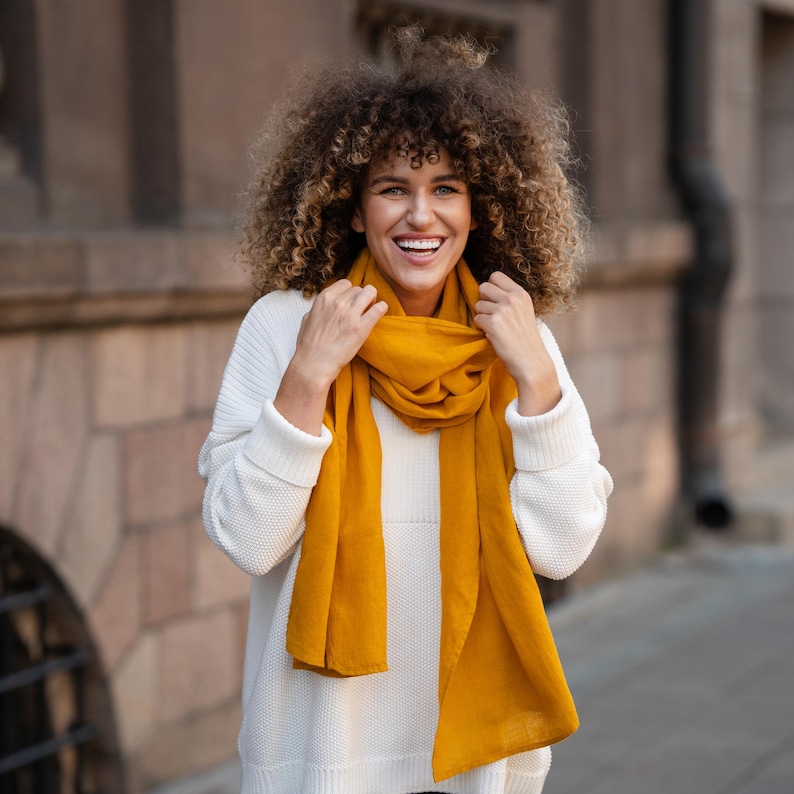 May include: A woman with curly brown hair is smiling and wearing a white sweater with a long, mustard yellow scarf.