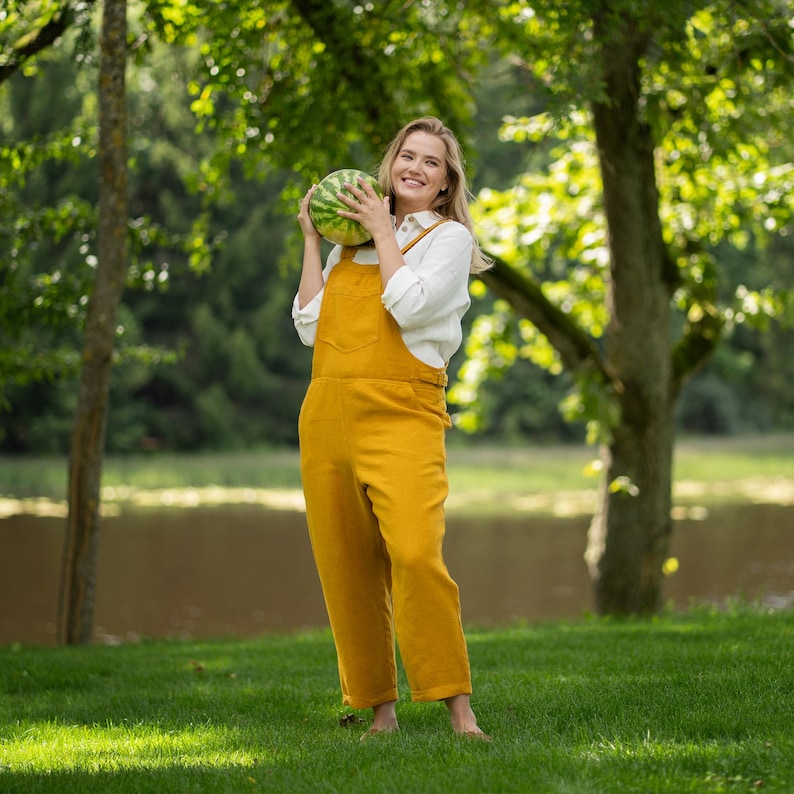 A cheerful, barefoot woman stands on the grass near a serene lake, playfully holding a small whole watermelon up to her shoulder. Her summery outfit features a pure white collared linen shirt worn underneath a bright, spicy yellow linen jumpsuit.