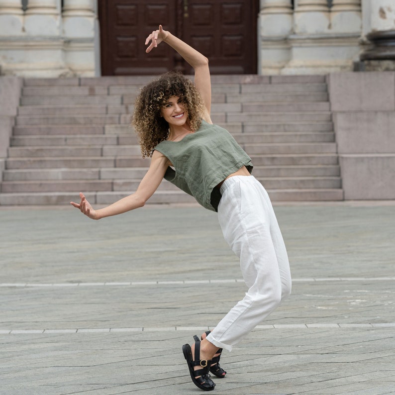 The model is wearing a linen clothing set, which includes a sleeveless crop top in stone green color and white high-waisted pants with pockets. She is standing on her toes, with her hands in the air,her legs bent at the knees.