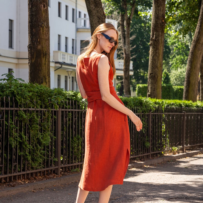 May include: A woman wearing a rust-colored sleeveless linen dress with a belt. She is wearing sunglasses and standing in front of a fence.