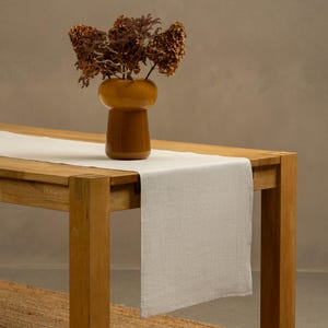 The menique natural color linen table runner on top of a wooden table. There is a vase on top of the runner.