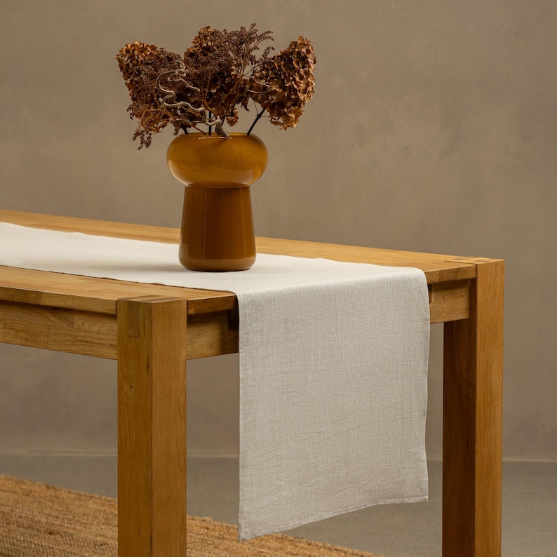 The menique natural color linen table runner on top of a wooden table. There is a vase on top of the runner.
