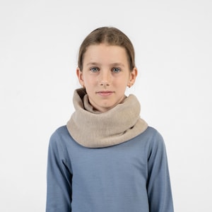 Può includere: Uno scaldacollo in maglia beige indossato intorno al collo, caratterizzato da un tessuto morbido e testurizzato con motivo a coste. La sciarpa è abbinata a una maglia a maniche lunghe blu tinta unita, creando un contrasto neutro con il capo base.
