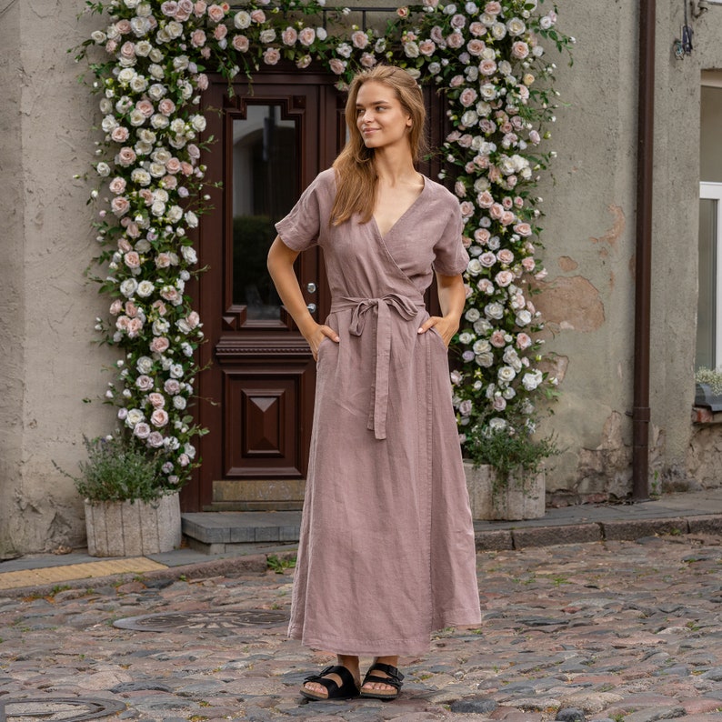 The model wears the ELEANOR maxi wrap dress in faded rose. She is standing full height, with her hands in the side pockets. The wrap design features a v-neck and a belt around the waist.