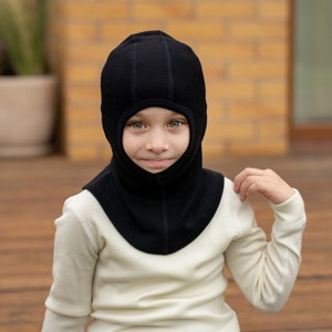 May include: A child wearing a black fleece balaclava with a white turtleneck shirt.