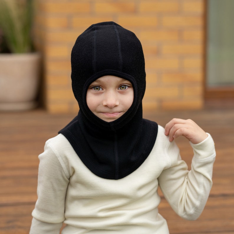 May include: A child wearing a black fleece balaclava with a white turtleneck shirt.
