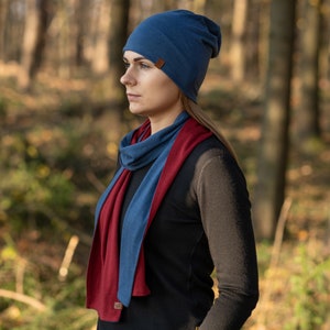 May include: A blue beanie with a brown leather tag and a matching blue and burgundy scarf. The scarf is folded in half and worn around the neck.