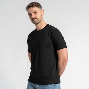May include: A black crew neck t-shirt with short sleeves. The shirt is made of a soft, textured fabric. The model is wearing blue jeans. The shirt has a small brown tag on the bottom right side.