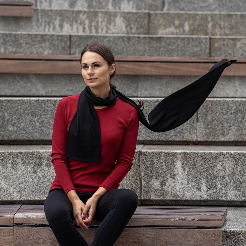 May include: A woman wearing a red long-sleeved shirt and black pants sits on a wooden bench. She is wearing a black scarf that is flowing in the wind.